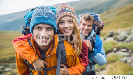 Three cheerful hikers, equipped with backpacks and warm hats, pose closely with bright smiles against a scenic outdoor backdrop. This image captures the camaraderie and happiness of friends enjoying 129713570