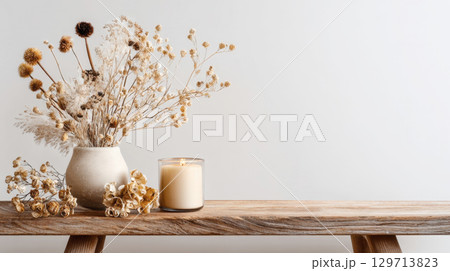 This calming still life showcases a textured ceramic vase filled with natural dried flowers and grasses, alongside a lit candle, all resting on a rustic wooden surface against a clean white background 129713823