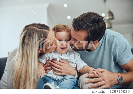 Mom, dad and baby kiss on sofa in living room of happy family home, young parents and child together. Love, happiness and couple kissing kid with smile while sitting on couch in New York apartment. 129715029