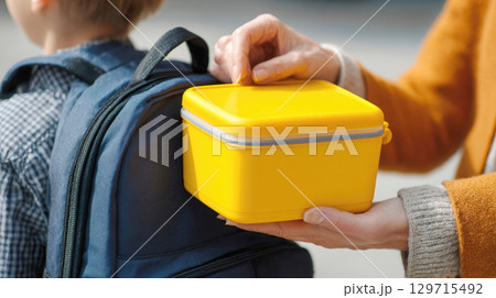 A nurturing moment as a parent places a bright yellow lunchbox into their child's backpack, symbolizing care, healthy habits, and preparation for a new school day and future success 129715492