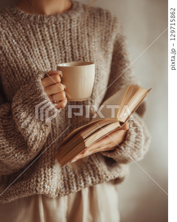 A cozy, warm image of a person wearing a chunky knit sweater, holding a warm cup and an open book, creating a serene and comfortable atmosphere perfect for a quiet moment of relaxation 129715862