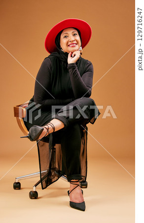 Fashion, fancy and a mature model woman in studio on a brown background sitting on a chair for trendy clothes. Portrait, hat and clothing with an attractive senior female seated to promote style 129716247