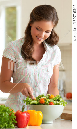 A happy woman smiles while tossing fresh, colorful salad in large bowl in a sunlit kitchen. Themes of joy of healthy cooking, with focus on fresh ingredients like bell peppers, tomatoes, leafy greens 129716345
