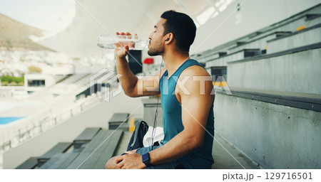 Stadium, man and drink water with earphones for fitness, hydration and running break. Outdoor, athlete person and relax with liquid beverage for training podcast, exercise and music for workout rest 129716501