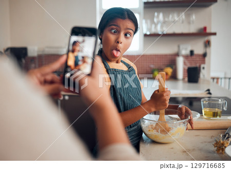 Funny child, phone and baking in home with mother taking photo for social media or food blog post in their home kitchen while making cake. Girl kid making comic face while cooking with a woman Funny child, phone and baking in home with mother taking photo for social media or food blog post in their home kitchen while making cake. Girl kid making comic face while cooking with a woman 129716685