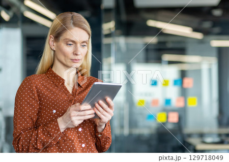 A focused businesswoman in an office setting, holding a tablet, appears engrossed in her work. The modern office has glass walls. 129718049