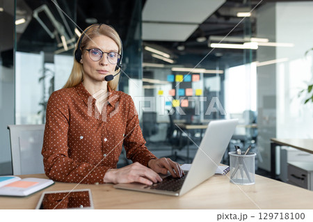 A blonde woman with glasses wearing a headset works at her desk on a laptop in a modern, bright office setting. A blonde woman with glasses wearing a headset works at her desk on a laptop in a modern, bright office setting. 129718100