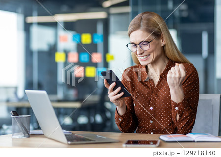Smiling businesswoman wearing glasses celebrates exciting news on smartphone in office. Enthusiastic reaction highlights professional success and communication. Smiling businesswoman wearing glasses celebrates exciting news on smartphone in office. Enthusiastic reaction highlights professional success and communication. 129718130