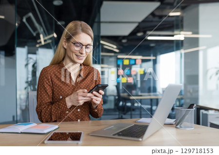 Joyful businesswoman using phone while sitting inside office, woman typing message using app on smartphone, working with laptop inside office. Joyful businesswoman using phone while sitting inside office, woman typing message using app on smartphone, working with laptop inside office. 129718153