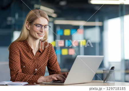 Successful joyful woman working with laptop inside office at workplace. Businesswoman typing on keyboard, satisfied with results of achievement, mature company employee. Successful joyful woman working with laptop inside office at workplace. Businesswoman typing on keyboard, satisfied with results of achievement, mature company employee. 129718185