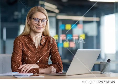 Portrait of a successful businesswoman at her workplace inside the office. Office worker smiling and looking at the camera, sitting at a desk with a laptop. 129718189