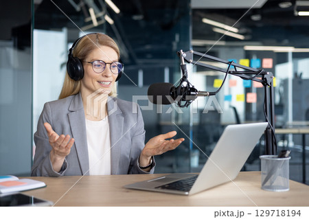 Mature woman businesswoman in a modern office setting, using headphones and a microphone for podcast production. The image portrays professionalism and highlights a technological workspace. Mature woman businesswoman in a modern office setting, using headphones and a microphone for podcast production. The image portrays professionalism and highlights a technological workspace. 129718194