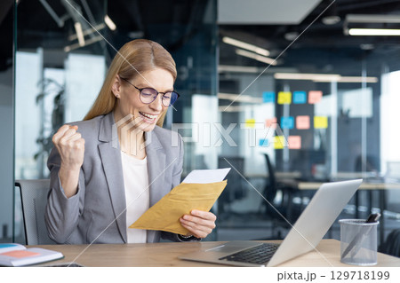 Mature businesswoman smiles with joy holding an envelope using laptop. Representing professional success and achievement, this image captures a moment of accomplishment in office environment. 129718199
