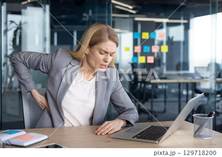 A mature businesswoman is sitting at her desk in discomfort, experiencing back pain. This image highlights health issues such as poor posture or prolonged sitting in a professional environment. 129718200