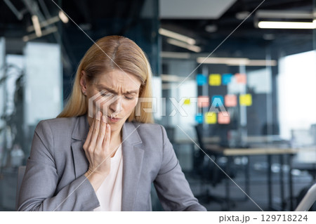 Mature woman businesswoman holding her jaw in pain at work in an office. Depicting discomfort possibly from a dental issue, this conveys feelings of distress and the need for healthcare attention. Mature woman businesswoman holding her jaw in pain at work in an office. Depicting discomfort possibly from a dental issue, this conveys feelings of distress and the need for healthcare attention. 129718224