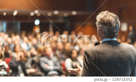 A professional public speaker is holding a microphone and addressing a large crowd in a conference hall. The image conveys leadership, communication, and inspiration in a professional business setting A professional public speaker is holding a microphone and addressing a large crowd in a conference hall. The image conveys leadership, communication, and inspiration in a professional business setting 129718352