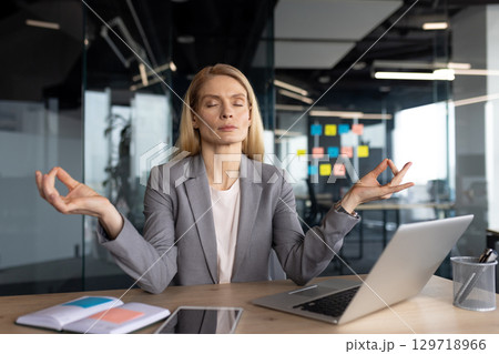 Mature businesswoman seated at her desk performing mindfulness meditation to relax and focus. Maintaining a calm and composed expression, she sits surrounded by work materials Mature businesswoman seated at her desk performing mindfulness meditation to relax and focus. Maintaining a calm and composed expression, she sits surrounded by work materials 129718966
