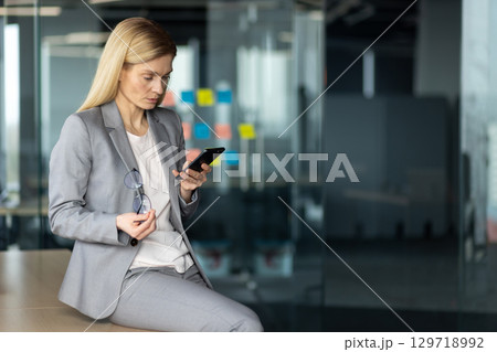 Mature businesswoman in gray suit using smartphone in modern office setting. Holding glasses, she focuses on device, portraying leadership, multitasking, and professionalism in corporate environment. 129718992