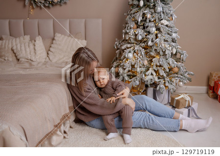Young mother holding baby near decorated Christmas tree in cozy bedroom, front view 129719119