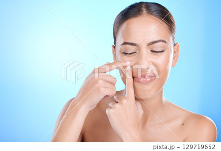 Woman, skincare and squeeze pimple on face with blue studio background. Skin care, morning facial and young model with flaws, blemish and imperfection pop acne or blackhead with fingers and hands 129719652