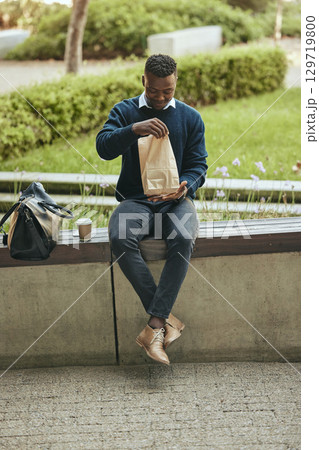 Black business man on a lunch break in the city, sitting and eating a meal or sandwich in a brown paper bag. Hungry worker relaxing with a tasty snack, food on the go for a professional entrepreneur 129719800