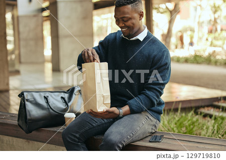 Office employee with a coffee, a paper sandwich bag and a smile, on a bench for a break from his busy work schedule. Happy, hungry businessman, time to eat, with his food and drink outside for lunch. 129719810