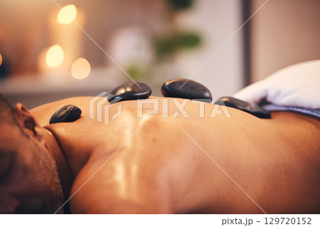 Massage, hot stone and relax with a man in a spa, lying on a table or bed for physical therapy at a luxury resort. Wellness, rock and zen with a male customer relaxing in a health center for rest 129720152