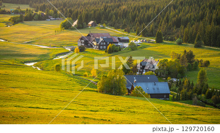 Evening light casts a warm glow over Jizerka Village in the Jizera Mountains. The tranquil landscape features fields, forests, and quaint houses surrounded by nature's beauty. 129720160