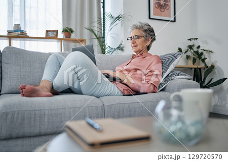 Senior woman, laptop and relax on sofa in the living room checking email, typing or writing at home. Elderly female freelancer or writer relaxing on lounge couch working or reading on computer 129720570