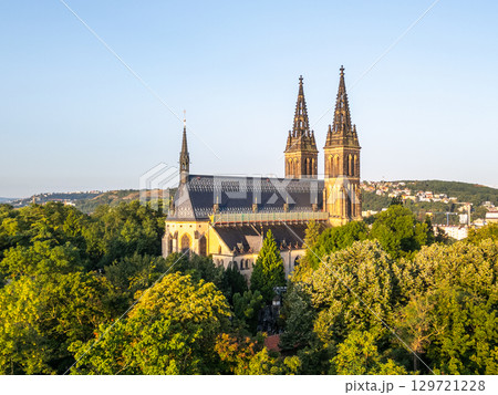Morning sunlight bathes the Vysehrad Basilica of St. Peter and St. Paul in warm hues, highlighting its impressive Gothic architecture amid lush greenery in Prague. 129721228