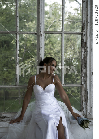 beautiful bride in a long elegant dress with a bouquet of lilies of the valley sits near the window 129723829