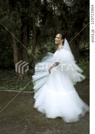 A bride in a white dress and a long veil stands among the trees 129723904