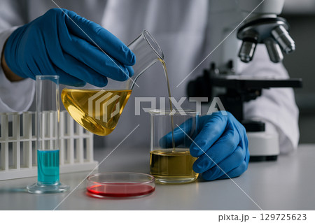 AI generated image of gloved hands pouring golden yellow liquid from an Erlenmeyer flask into a beaker with test tubes petri dishes and microscope in a clean professional laboratory 129725623