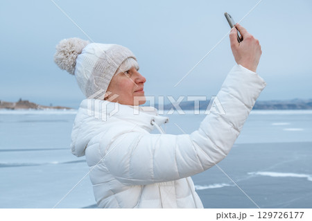 woman takes winter selfie by icy lake with snow covered landscape in background. closeup. 129726177