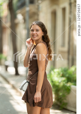 Back view of cute young girl wearing stylish short summer dress with white handbag walking along shady tree lined street enjoying spring day in Barcelona 129726857