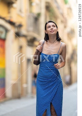 Cheerful brunette girl in blue dress walking along the street among architecture with bag 129726919