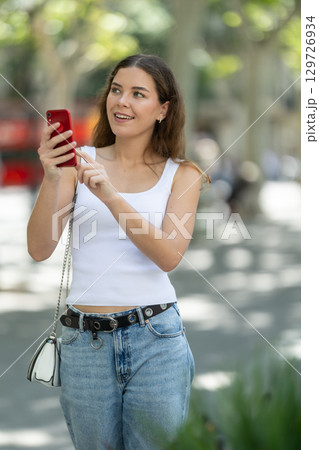 Happy woman looking to phone in hands and chatting in city 129726934