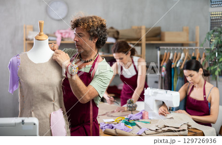Middle-aged male dressmaker working with mannequin while others sewing and drawing with curves Middle-aged male dressmaker working with mannequin while others sewing and drawing with curves 129726936