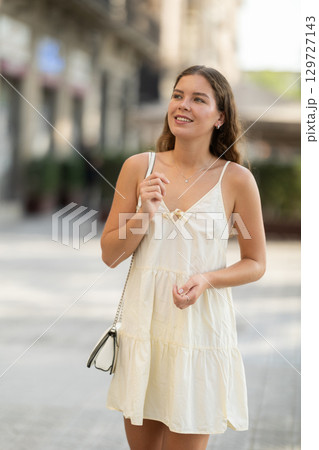Cheerful girl in white dress walking along the street among architecture with bag 129727143