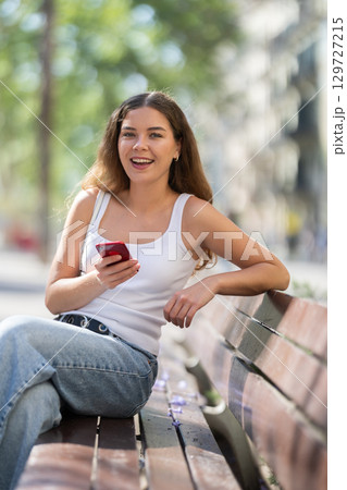 Portrait of happy delightful girl with mobile phone in hands, relaxing on bench in summer park Portrait of happy delightful girl with mobile phone in hands, relaxing on bench in summer park 129727215