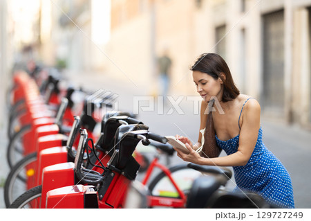 Brunette in blue dress rents electric bike - pays with mobile phone at street parking 129727249