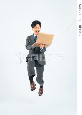 ノートパソコンを持ってジャンプする若いビジネスマンの全身写真 129727369