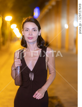 Young woman walking along warmly lit urban pathway of Barcelona at night 129727649