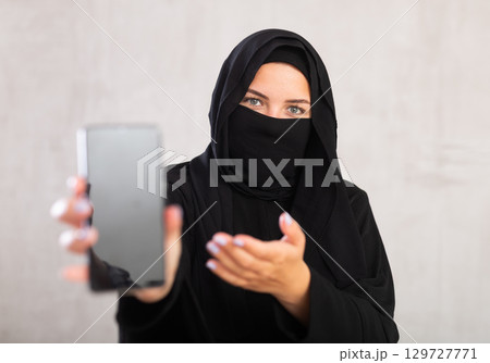 young Muslim girl with her face covered by burka shows empty dark smartphone screen. young Muslim girl with her face covered by burka shows empty dark smartphone screen. 129727771