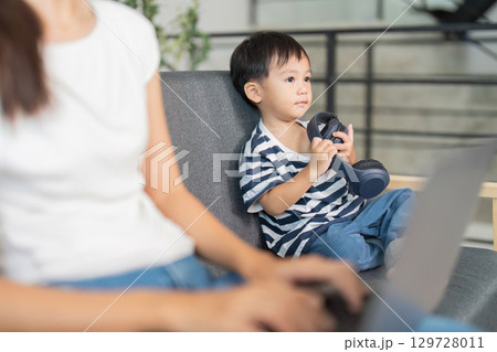 Child holding headphones while mother works on laptop at home Child holding headphones while mother works on laptop at home 129728011