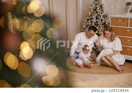 Young family with baby playing near Christmas tree in cozy room with white wall and wooden floor Young family with baby playing near Christmas tree in cozy room with white wall and wooden floor 129728934