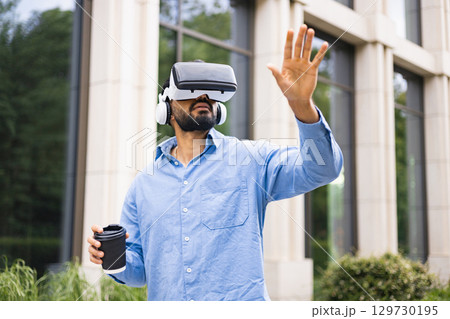 A man wearing a VR headset and headphones, reaching out while holding a coffee cup outdoors near a building. 129730195