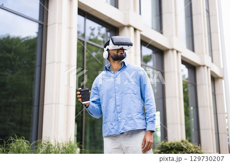A man wearing a VR headset and holding a coffee cup stands outside a modern building. 129730207