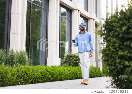 A man wearing a VR headset walks down a sidewalk, holding a coffee cup, in front of a modern building. A man wearing a VR headset walks down a sidewalk, holding a coffee cup, in front of a modern building. 129730211