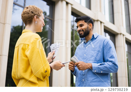 A woman hands money to a man outdoors, likely a financial transaction, against a building. A woman hands money to a man outdoors, likely a financial transaction, against a building. 129730219